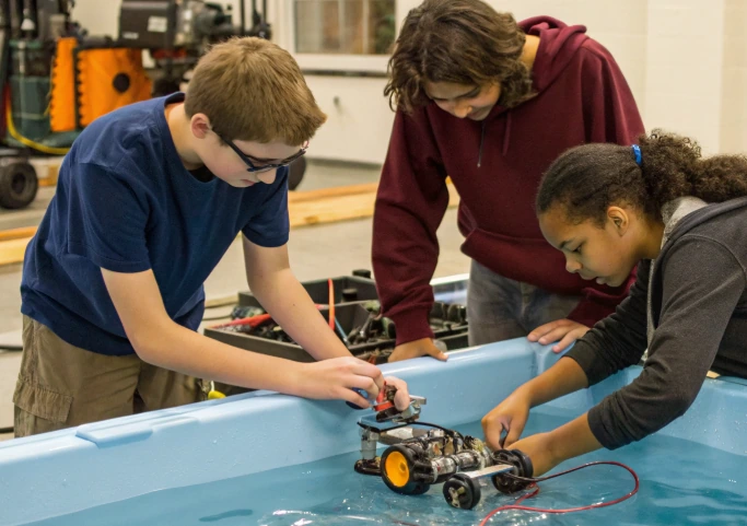 Three students testing an underwater robot in a water tank, showing why the underwater environment matters pedagogically for engineering trade-offs

